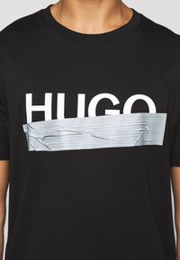 Osoba v černém tričku s bílým nápisem "HUGO", částečně zakrytým vodorovným pruhem stříbrné izolační pásky přes hruď.