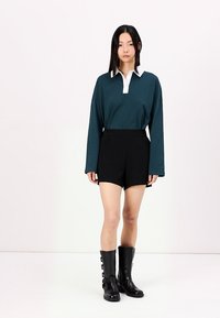 Zelena polo majica s dolgimi rokavi in belim ovratnikom, v kombinaciji s črnimi visokimi hlačami in črnimi škornji do kolen z naramnicami in zaponkami.