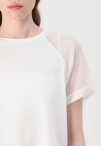 T-shirt bianco con maniche in tessuto leggero e trasparente, scollatura rotonda e texture morbida. Presenta una vestibilità comoda e un dettaglio di cucitura sottile.
