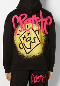 Sudadera negra y pantalones con texto en estilo graffiti de color rosa brillante que dice "CREATED" y un cuadrado amarillo que presenta un diseño de cara abstracta negra en la parte trasera.