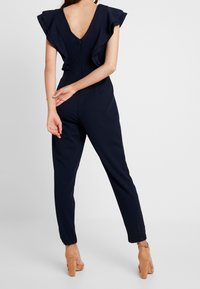 Náměsíčný modrý overal s volány na rukávech, hlubokým V výstřihem na zádech, přiléhavým střihem, zúženými nohavicemi a zipovým uzávěrem na zádech. Hladká textilie.