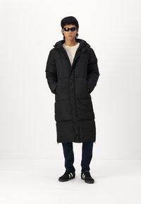 Zwarte, lange pufferjas met een capuchon, ritssluiting en twee zakken aan de voorkant. Draag het over een lichtgekleurde shirt en donkere jeans, met zwarte sneakers.