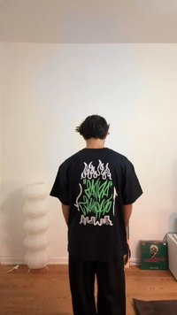 T-shirt preta oversized com texto de graffiti verde e desenhos de chamas brancas nas costas. Usada com calças pretas largas.