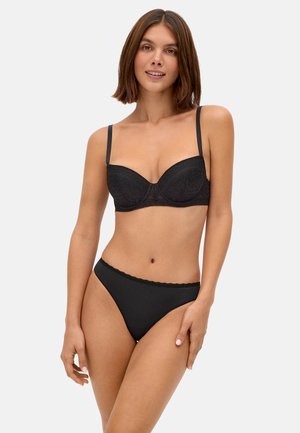 Mujer con cabello castaño hasta los hombros, vestida con un sujetador de encaje negro y bragas negras a juego, posando frente a un fondo claro.