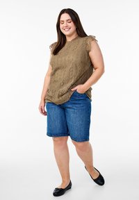 Olivi vihreä hihaton pitsipaita, jossa on frillikoristeita, yhdistettynä vaaleansinsisiin denim Bermuda-shortsit. Puettuna mustilla tasapohjaisilla kengillä, seisoo tasaisella taustalla.
