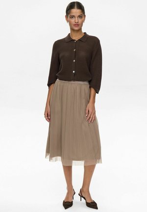 Brown ribbed-knit blouse with buttons, paired with a pleated beige skirt. The outfit features elegant flow and texture. Brown high-heeled shoes complete the look.