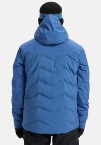 Person mit einer blauen gesteppte Winterjacke mit Kapuze, schwarzen Handschuhen und einer schwarzen Mütze mit blauem Band, die sich abwendet.