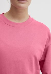 Lys pink sweatshirt med en rund halsudskæring, ribbede manchetter og en blød tekstur, der viser en del af overkroppen fra brystet og op.