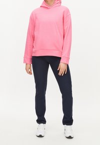 Růžová mikina s kapucí a dlouhými rukávy, s přední kapsou, v kombinaci s tmavě modrými kalhotami. Bílé tenisky dotvářejí celý outfit.