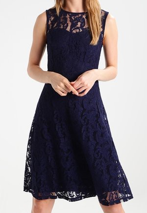 Laivastonsininen pitsimekko, jossa on istuva yläosa ja liehuva helma. Mekossa on koristeellinen pitsikerros ja pyöreä pääntie.