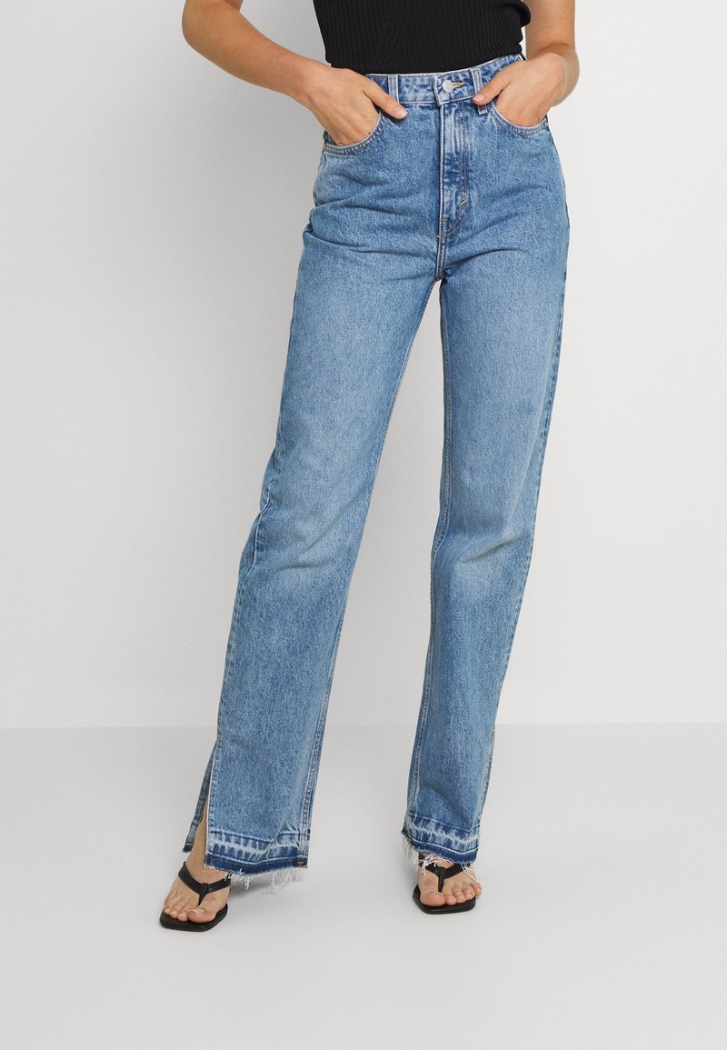 Personne portant un jean bleu taille haute avec des fentes aux chevilles et des ourlets effilochés, associé à des sandales noires à brides et un haut noir sans manches.