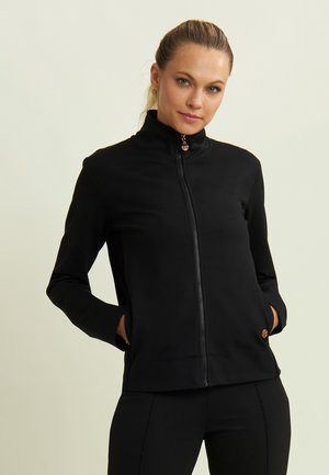 Veste noire à fermeture éclair avec un col haut, dotée de poches latérales et d'un accent en bronze sur la fermeture. Tissu lisse et extensible.