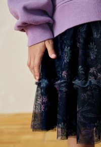 Egy gyermek keze egy virágmintás fekete tüll szoknya mellett pihen, lavanda színű, hosszú ujjú pulóvert viselve, fa padlón állva.