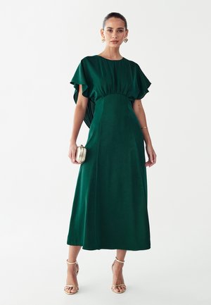 Zelena obleka srednje dolžine z nabranim pasom, krilci na rokavih in gladko teksturo. Povezana z majhno kovinsko torbico in sandali s trakovi.