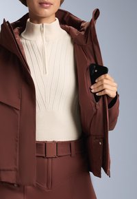 Barna szigetelt dzseki nyitva pihen, felfedve egy krémszínű bordás pul�óvert. Egy fekete telefon részben látható a dzseki zsebében. Az öv a dzseki színével megegyező színű.