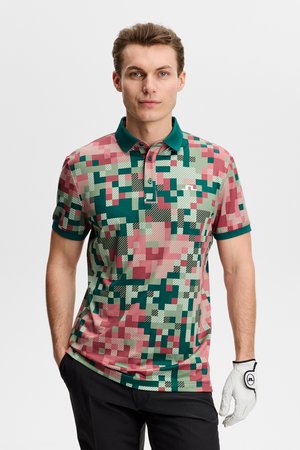 Mann iført en pikselert camouflagemønstret poloskjorte i grønt og rosa, svarte bukser og en hvit golfhanske på høyre hånd.