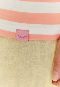 Ružovo-biely pruhovaný bavlnený golier s ružovou etiketu zobrazujúcou grafiku rias, v kombinácii so svetložltým textúrovaným kusom oblečenia.