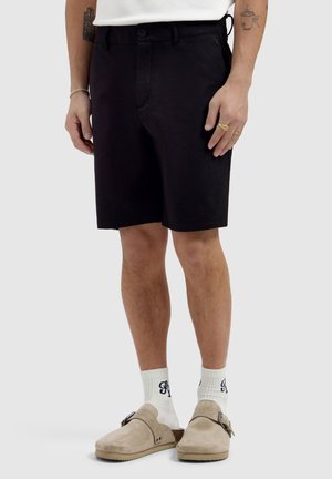 Mann trägt schwarze, knielange Shorts, weiße Socken mit einem Logo, beige Slipper sowie sichtbare Arm-Tattoos und Ringe.