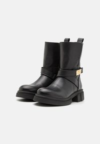 Bottines noires en cuir avec un talon épais, une large sangle et des accessoires en métal doré. Comprend des languettes pour un enfilage facile. Texture lisse.