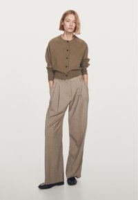 Brown knitted cardigan with black buttons, paired with light beige wide-leg trousers. Model stands with hands in pockets, wearing black flats.