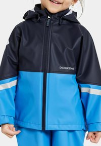 Vattentät jacka med färgblock i marinblått och ljusblått, dragkedja, huva, reflexdetaljer, slät glansig yta, logotyp på bröstet.