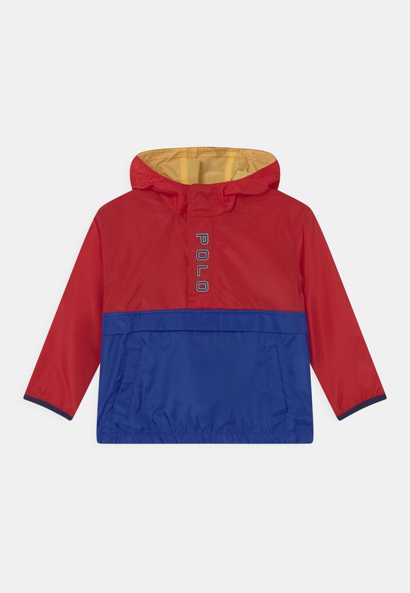 Červená a modrá větrovka se žlutou kapucí, s přední kapsou a logem „POLO“. Hladký povrch a elastické manžety.