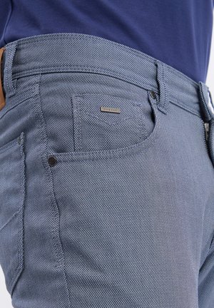 Zbliżenie na niebieską teksturowaną kieszeń spodni z małą metalową metką marki i widoczną szlufką na pasek, noszone z granatową koszulą.