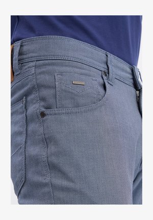 Zbliżenie na niebieską teksturowaną kieszeń spodni z małą metalową metką marki i widoczną szlufką na pasek, noszone z granatową koszulą.