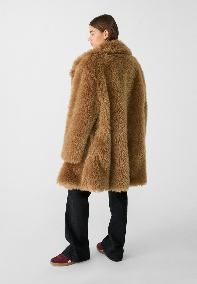 Cappotto in pelliccia sintetica marrone con un ampio colletto, taglio oversize e superficie testurizzata, abbinato a jeans scuri e sneaker bordeaux.