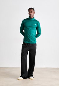 Maglietta a maniche lunghe zip-up color turchese con colletto alto e logo. Abbinata a pantaloni ampi neri e calzature chiare. Tessuto dalla trama liscia.