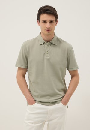 Jeune homme debout les mains dans les poches, portant un polo à manches courtes vert clair et un pantalon blanc sur un fond neutre.