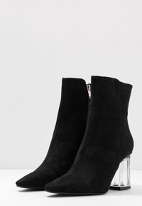 Botas de tobillo de ante negro con punta afilada y tacón de bloque de acrílico transparente. Cuenta con una cremallera lateral para el cierre. Textura suave en todo.