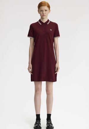 Femme debout portant une robe polo bordeaux avec des bordures blanches au col et aux manches, des chaussettes noires et des mocassins noirs, sur un fond uni.