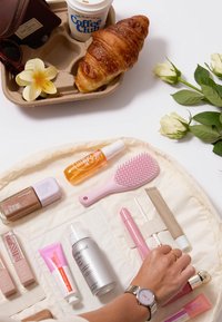 Organizer cosmetico con vari prodotti di bellezza. Include un pennello rosa, più tubetti, una lattina spray argentata e una bottiglia di fondotinta marrone.