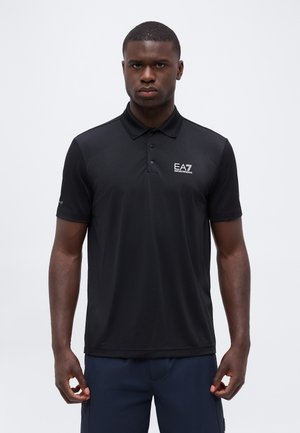 Mand iført en sort EA7 Emporio Armani poloshirt og marineblå bukser, stående mod en ensfarvet lys baggrund.