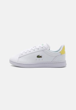 Hvid lavtop sneaker med hvide snørebånd, en grøn krokodille-logo på siden og en gul hælflap.