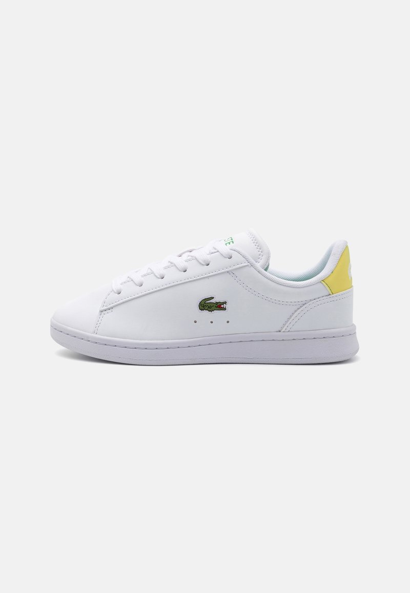 Baskets basses blanches avec lacets blancs, un logo crocodile vert sur le côté et une languette jaune au talon.