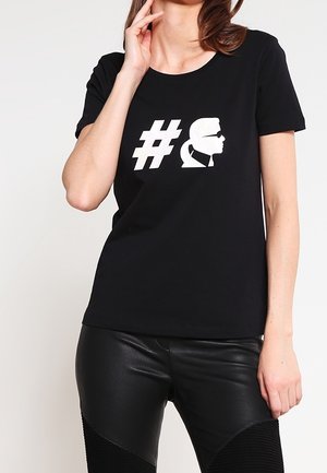 Vrouw draagt een zwart T-shirt met een wit hashtag-symbool en een zijprofiel silhouet van een persoon met zonnebril erop gedrukt.