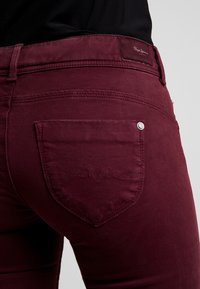 Burgundy jeans feature a slim fit, a back pocket with wavy stitching detail, and a logo patch at the waistband. Soft, textured fabric.