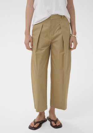 Személy bézs színű, bő, rövidített nadrágot, fehér felsőt, ezüst karkötőt és fekete, négyzet alakú orrú flip-flop papucsot visel, tiszta háttér előtt áll.