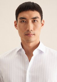 Jasnoniebieska koszula w białe paski z kołnierzykiem, zapinana na guziki i o miękkiej fakturze. Zbliżenie na twarz mężczyzny modela.