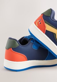 Baskets colorées avec un mélange de matériaux synthétiques bleu marine, orange, vert et jaune. Visage souriant distinctif sur le côté, semelle en caoutchouc.