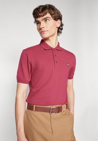 Bordový polo tričko s krátkym rukávom, golierom a prietkom na tri gombíky; na ľavej hrudi sa nachádza malá žltá emblema.