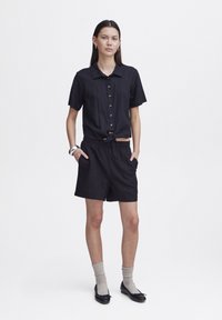 Camicia nera in lino a maniche corte con chiusura a bottoni e nodo frontale, abbinata a pantaloni corti neri, calze grigie e ballerine nere. Taglio semplice e rilassato.