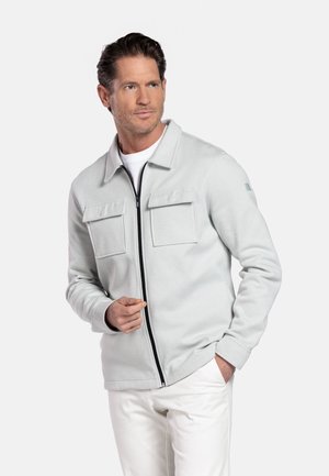 Man wearing light gray zip-up jacket with two front flap pockets over white shirt and white pants, looking to the side.