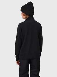 Persona che indossa un berretto nero, una giacca in fleece nera e pantaloni neri, girata di spalle contro uno sfondo grigio chiaro uniforme.