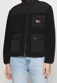 Czarna polarowa kurtka z zamkiem błyskawicznym z przodu, dwoma bocznymi kieszeniami na zamek oraz kieszenią naszywaną na piersi. Posiada stójkę i fakturowany materiał.