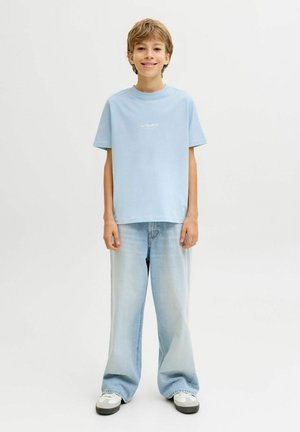 Hellblaues Baumwoll-T-Shirt mit rundem Ausschnitt und kurzen Ärmeln, kombiniert mit hellblauen Jeans mit weitem Bein. Weiße Sneaker mit schwarzen Akzenten.