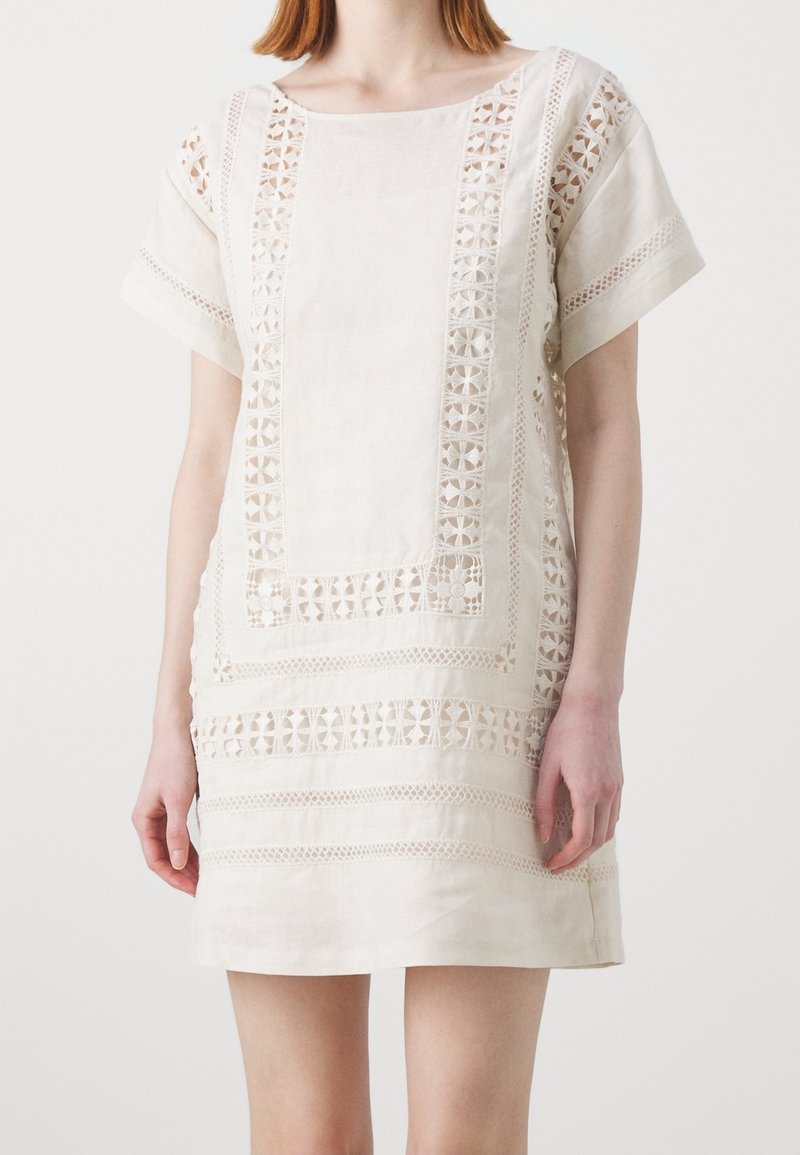 Femme portant une robe crème à manches courtes et longueur genoux, avec des panneaux en dentelle florale et des motifs géométriques ajourés sur le devant et les manches.