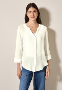 Hvid knappet blouse med en tekstureret, krøllet stof, der har en rund halsudskæring, trekvartærmer og en buet kant.